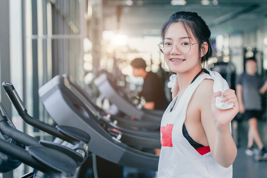 Asian teenage braces girl cardio training exercise in sport club with towel on her shoulders smiling after workout on treadmill in the gym looking camera.