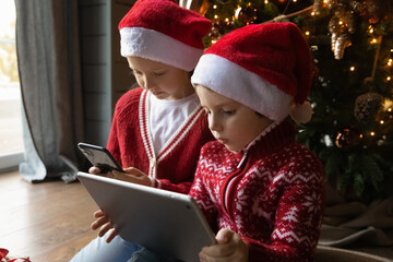Small Caucasian boy and girl children look at gadget screens play computer online games on winter holidays at home. Little brother and sister kids engaged in digital virtual activity on devices.