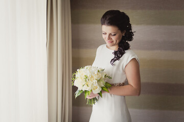 Pretty young Bride in simple wedding dress with her wedding bouquet. Black-haired woman with wedding hair-style with a long tail. Boudoir morning of the bride in modern room