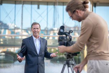 Long-haired operator filming smiling mature male reporter in front of fountain
