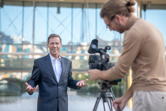 Long-haired operator filming smiling mature male reporter in front of fountain