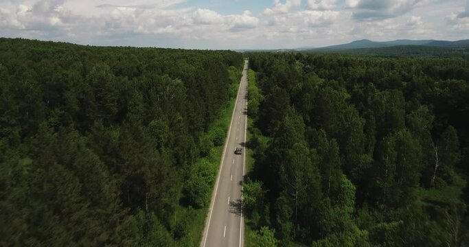 One dark car driving alone / travel on empty straight asphalt road freeway through dense forest corridor at summer sunny day. Aerial drone wide view.