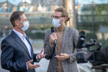Fair-haired male journalist interviewing businessman in front of camera, both wearing masks