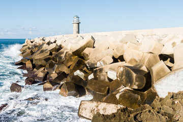 Wave crashing against concrete tetrapods wave breakers with a small lighthouse in the background in...