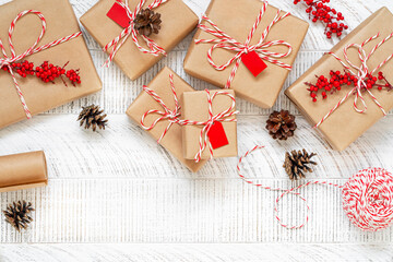 Top view of decorated boxes on the white wooden background. Preparing christmas gifts with craft paper, striped twine string, branches with red berries and pine cones. New year flat lay. Copy space.