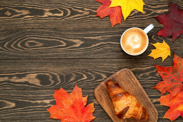 Obraz premium A cup of coffee, homemade croissant and colorful maple leaves on the wooden background. Autumn flat lay with copy space. Good morning breakfast.