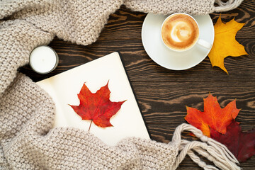 Cozy and warm autumn flat lay with a cup of coffee, open book, candle, knitted plaid, red, orange and yellow leaves on the wooden background. 