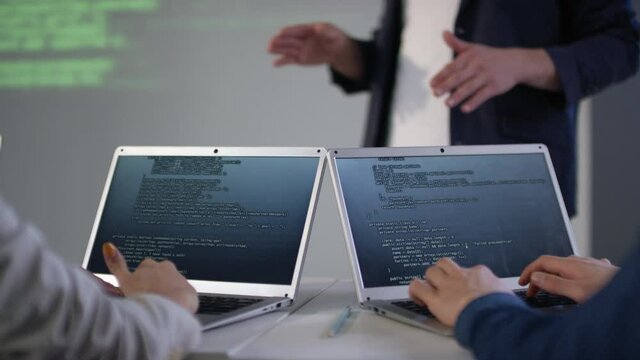 Panning shot of classmates typing codes on laptops while listening to male computer programming tutor explaining topic in background