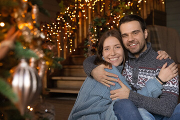 Fototapeta premium Portrait of happy young couple pose near decorated Christmas tree hugging cuddling celebrating winter holidays together. Smiling man and woman enjoy New Year celebration at home together.