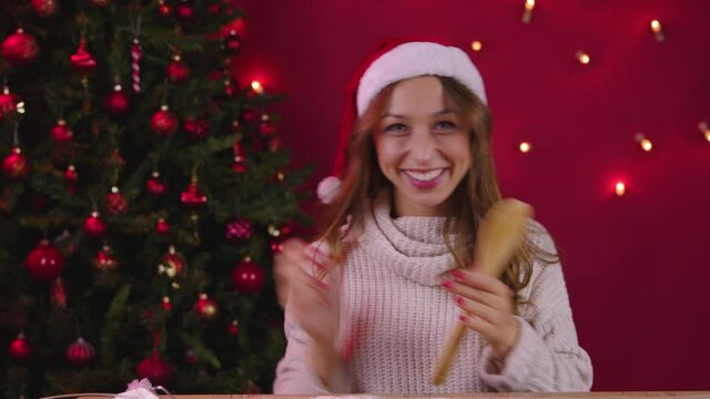 Cute long-haired girl singing using wooden spatula instead of microphone on Xmas decoration with Christmas tree. Christmas bakery, Good festive mood concept. Merry Christmas and Happy New Year