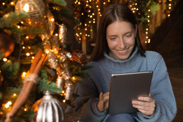 Smiling young Caucasian woman sit near decorated Christmas tree have fun browsing internet on...