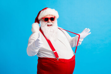 Fototapeta na wymiar Photo of modern style santa claus with big abdomen pull suspender point index finger gloves x-mas christmas noel ads wear sunglass cap overalls pants isolated blue color background