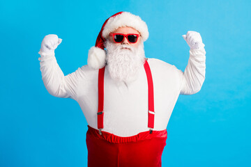 Fototapeta na wymiar Photo of confident santa claus with big stomach show hands muscles x-mas christmas fairy magic strength wear headwear sunglass overalls suspender isolated over blue color background