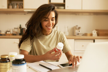 Biohacking and superfoods concept. Cheerful young Latin female nutritionist holding bottle of dietary supplements for muscle building. Pretty girl using laptop ordering vitamins via online store - obrazy, fototapety, plakaty