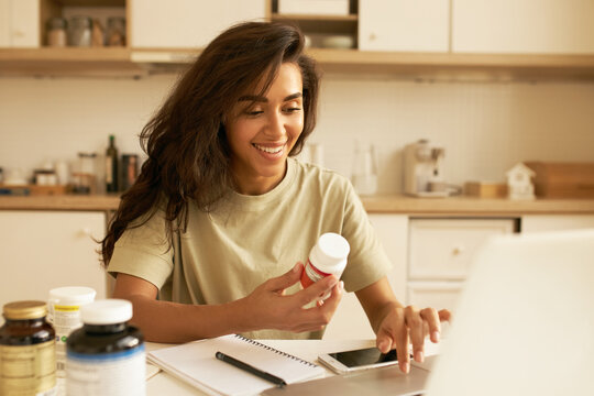Biohacking and superfoods concept. Cheerful young Latin female nutritionist holding bottle of dietary supplements for muscle building. Pretty girl using laptop ordering vitamins via online store