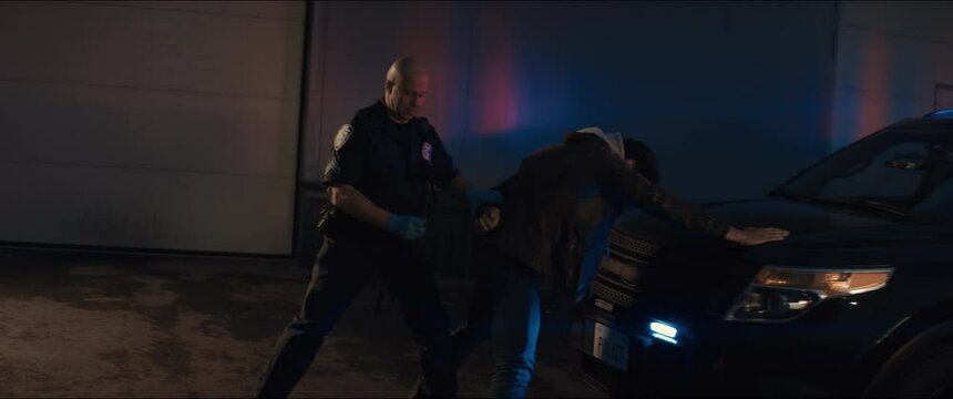 WIDE Police officer searches and handcuffs a suspect, African-American Black criminal. Police car lights flashing in the background. Shot with RED cinema camera and 2x Anamorphic lens