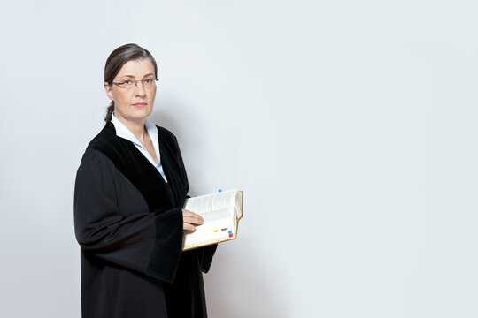 Middle aged female judge in black gown with a law book, neutral background, copy space.