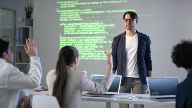 Young male it teacher discussing computer programming and program coding with teenage mixed race students with screen projector in background