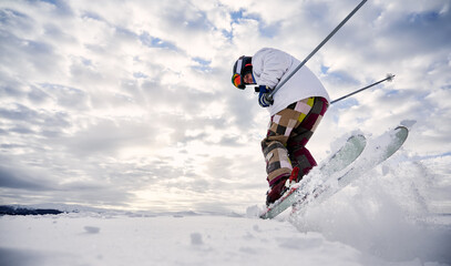 Fototapeta na wymiar Low angle view snapshot of skier's legs wearing colorful ski pants making a jump up on white snowy surface against beautiful cloudy sky. Copy space. Concept of winter sport activities.