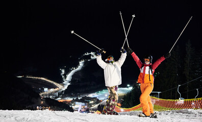 Cheerful male and female skiers in winter jackets and helmets raising ski poles. Joyful young man...