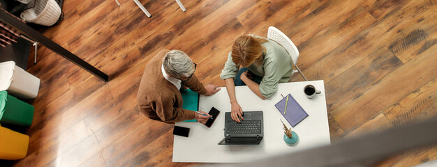 Two well-dressed female colleagues of different age discussing fashion having another colleague sitting at a table working with a PC