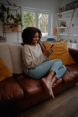 Obraz premium Beautiful african female watching video on smartphone at home while relaxing on couch