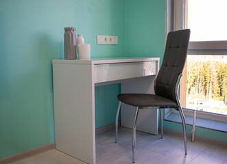 Wooden cosmetic table with vases and candle on green wall background. White table with soft chair in the corner of the room near big window.