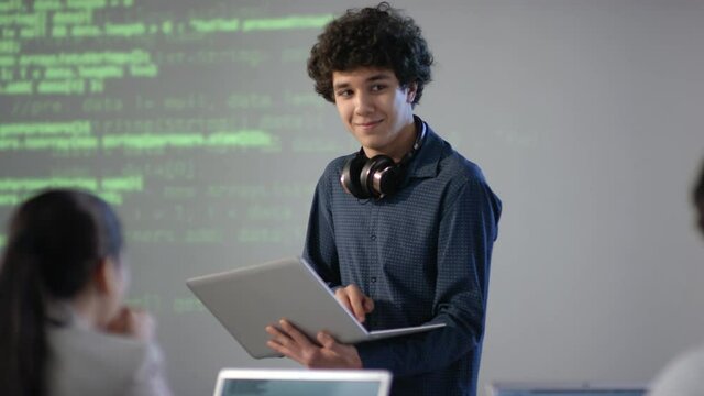 Medium closeup of cheerful mixed race student with laptop in hands making speech on computer programming topic smiling to classmates standing at board