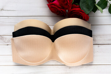 beige and black strapless nude bra on a wooden texture background next to rose flowers . Copy space for text input on health care and fitness. Balconette bra and lingerie for woman, top view.