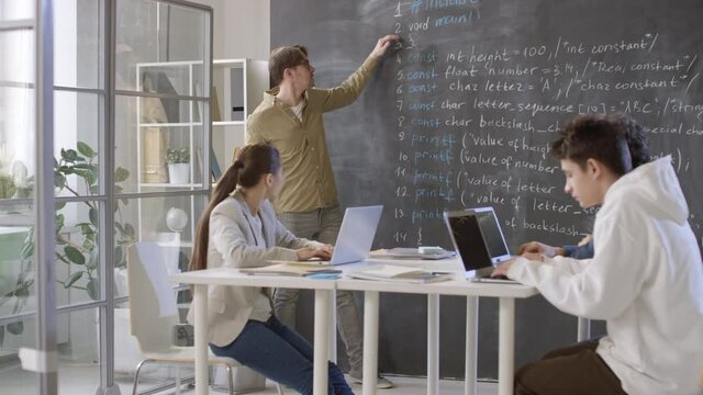 Sequence of shots of modern it professor in glasses explaining new material written on blackboard to team of teenage students sitting at desks with laptops typing something