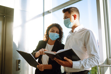 Two colleagues in protective face masks in modern office discussing together work issues standing near elevator. Business People back at work in office after quarantine. Covid-19.