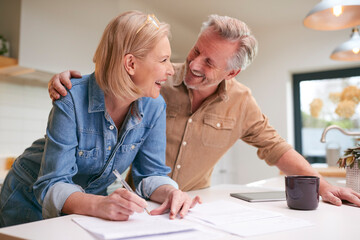 Mature Couple Reviewing And Signing Domestic Finances And Investment Paperwork In Kitchen At Home - obrazy, fototapety, plakaty