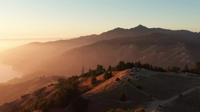 Aerial, campers on hilltop in Big Sur, California, camera pulls back to reveal the ocean below and the golden sunset.