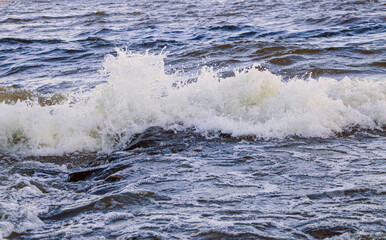 Naklejka premium waves run onto the shore and crash against the rocks, creating many splashes and splashes near the shore. river surf in stormy weather near a stone pebble coast with foamy splashing waves.