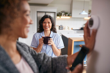 Same Sex Mature Female Couple Using App On Phone To Control Digital Central Heating Thermostat At...