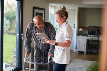 Senior Man In Dressing Gown Using Walking Frame Being Helped By Female Nurse With Digital Tablet