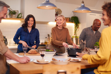 Group Of Mature Friends Meeting At Home Serving Food At Dinner Party And Drinking Wine Together