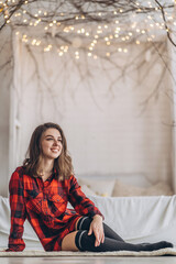 Fototapeta premium Pretty brunette girl sitting on the floor in red shirt and warm socks with christmas tree behind.