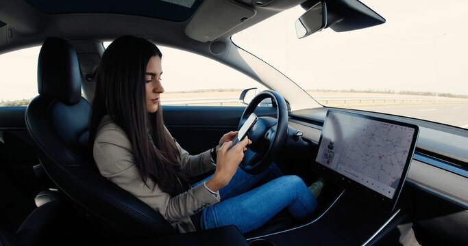 Portrait of beautiful young woman sitting in electric car and typing on smartphone. Happy Caucasian pretty female texting on cell phone in vehicle and smiling. Driverless car concept - Powered by Adobe