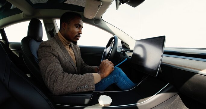 Young Concentrated Handsome Man In Electric Vehicle Tapping On Smartphone And Connecting Mobile Phone With Automated Car Handsome African American Male In Modern Auto Tapping On Dashboard Touch Screen