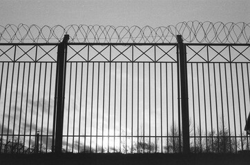 Silhouette of fence with barbed wire on film. Gloomy black and white overcast background with prison. Atmospheric scanned analog photography with grain and scratch. Lomography of private property.