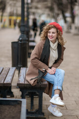 A cute girl in a red hat sits on a wooden bench on a city street. The girl smiles and looks to the side