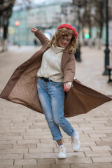 Beautiful brunette young woman in a red hat, jeans, and a coat happily whirls along the street