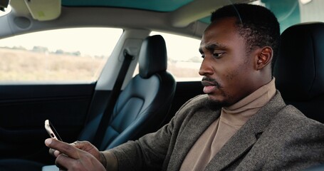 Close up portrait of African American man in electric car typing on smartphone and smiling to camera. Handsome happy male sitting in vehicle in good mood and texting on mobile phone. Transport concept