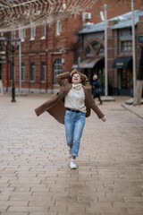 Beautiful brunette young woman in a red hat, jeans, and a coat happily runs down the street