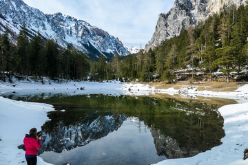 A woman standing next to a shore of Green Lake in Austrian Alps in winter and taking pictures of crystal clear water, great visibility. Reflections of Alps in calm lake's water. Winter wonderland.