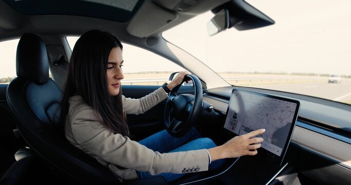 Beautiful Caucasian Young Woman Sitting In Electric Vehicle And Typing On Big Touch Screen. Cheerful Pretty Female Driver Tapping On-board Computer In Car And Searching Route On GPS Navigation Map