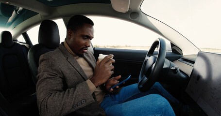 Side view of African American joyful man in self-driving electric car texting on smartphone while drinking coffee. Handsome young male traveling in modern autopilot vehicle and tapping on cellphone.