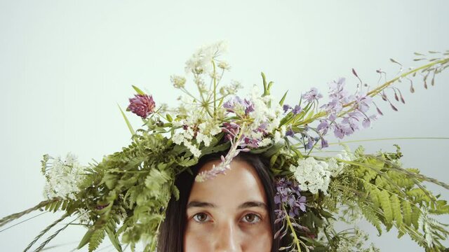 Close up shot of upper part of face of beautiful woman with flower wreath on her head looking at camera while posing in studio against white background