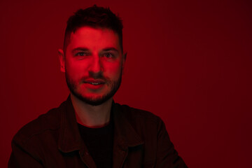 Low key portrait of bearded chubby young man looking at camera dressed casually in red light on dark red backdrop. UV ray skin damage